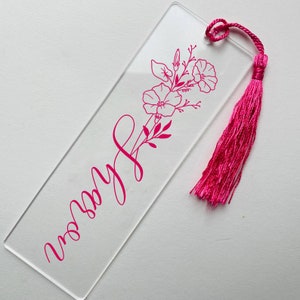 May include: A clear acrylic bookmark with a pink floral design and the word "worry" written in cursive. The bookmark has a pink tassel.
