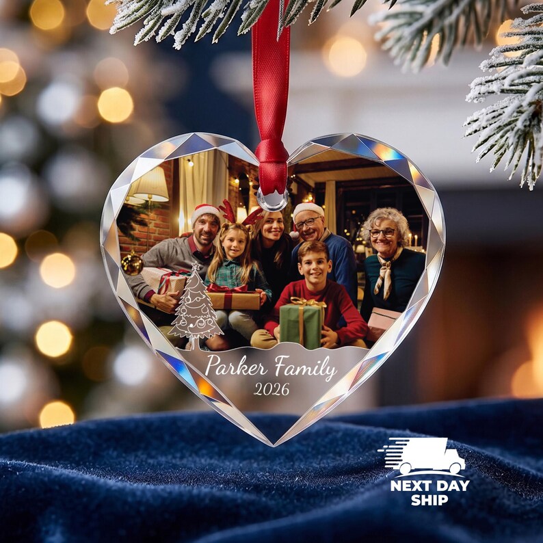 May include: A heart-shaped, faceted glass ornament featuring a printed family photograph, a white line-art Christmas tree, and the text "Parker Family 2026". The ornament hangs from a red satin ribbon against a blurred holiday background.