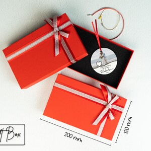 May include: Red gift box with a silver ribbon and bow. The box is 20 cm by 30 cm. Inside the box is a round, white ornament with a photo of a couple and the text "First Christmas as Mr. & Mrs. Robertson 2023".