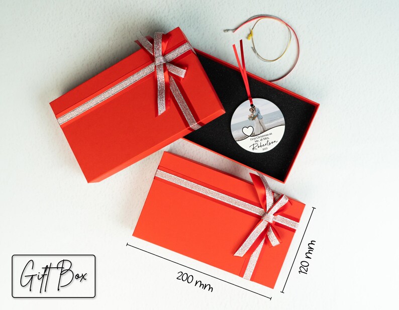 May include: Red gift box with a silver ribbon and bow. The box is 20 cm by 30 cm. Inside the box is a round, white ornament with a photo of a couple and the text "First Christmas as Mr. & Mrs. Robertson 2023".
