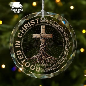 May include: A circular glass ornament featuring a wooden cross with roots extending downward, encircled by a crown of thorns. The text reads ROOTED IN CHRIST, ROOTED AND BUILT UP IN HIM AND ESTABLISHED IN THE FAITH AND OVERFLOWING WITH THANKFULNESS, COLOSSIANS 2:7.