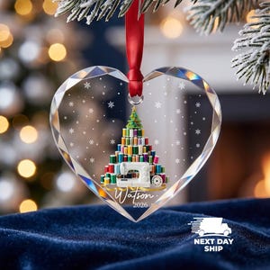 May include: A heart-shaped clear acrylic ornament with faceted edges, hanging from a red ribbon. The design features a Christmas tree made of colorful sewing thread spools, a white sewing machine, a yellow measuring tape, and the text Watson 2026.