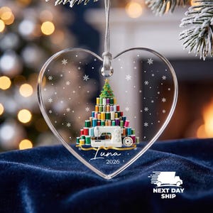 May include: A heart-shaped clear acrylic ornament featuring a printed graphic of a Christmas tree constructed from colorful sewing thread spools, a white sewing machine, and a yellow measuring tape. The text reads Luna 2026 with snowflakes.