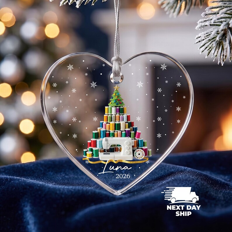 May include: A heart-shaped clear acrylic ornament featuring a printed graphic of a Christmas tree constructed from colorful sewing thread spools, a white sewing machine, and a yellow measuring tape. The text reads Luna 2026 with snowflakes.