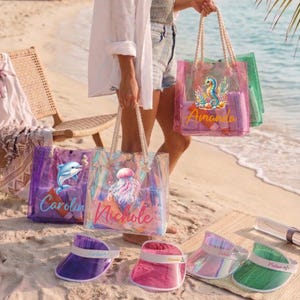 May include: Clear tote bags with rope handles, personalized with names and sea-themed designs. The bags are in various colors, including pink, purple, and green, and are filled with beach essentials. Visors in matching colors are also visible.