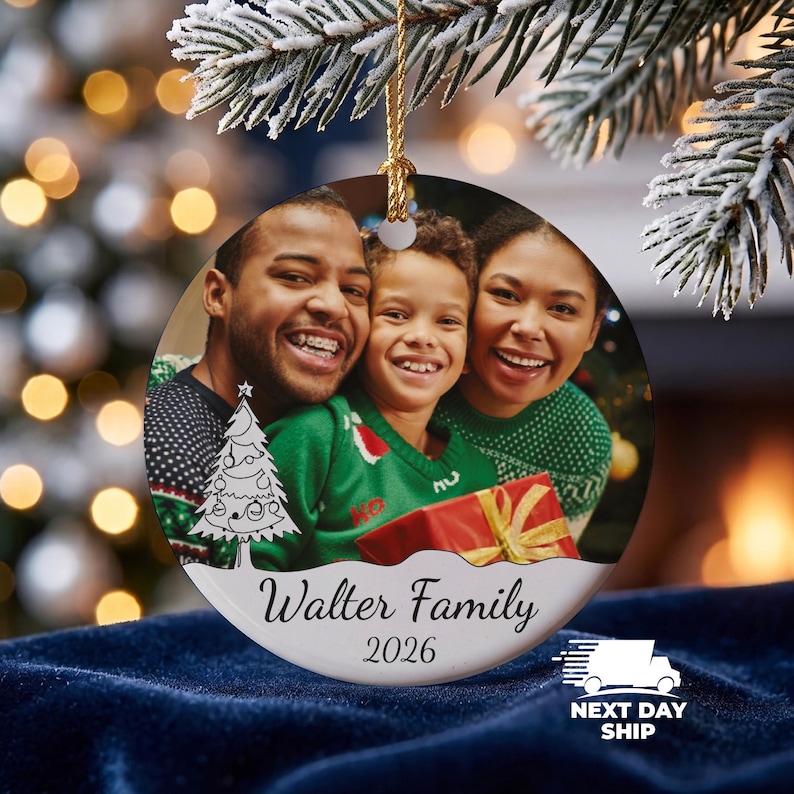 May include: A round ceramic Christmas ornament featuring a photograph of a family of three smiling. The ornament includes a line art illustration of a Christmas tree, the text Walter Family, and the year 2026 printed in black on a white base.