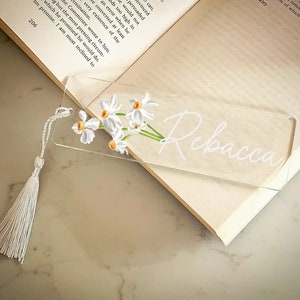 May include: A clear glass bookmark with a white tassel. The bookmark features a floral design with white flowers and yellow centers. The name "Rebecca" is written in white cursive on the bookmark.