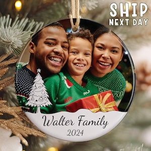 May include: A round ceramic Christmas ornament featuring a printed photograph of a family of three smiling. The ornament displays a white graphic of a Christmas tree, the text Walter Family, and the year 2024 printed in black script font.