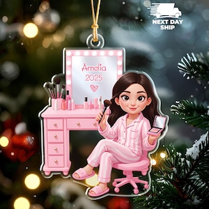 May include: A pink acrylic ornament featuring a cartoon illustration of a person in pink pajamas seated at a pink vanity table. The vanity has a mirror with the name "Amelia" and the year "2025". Makeup brushes and products are on the table.