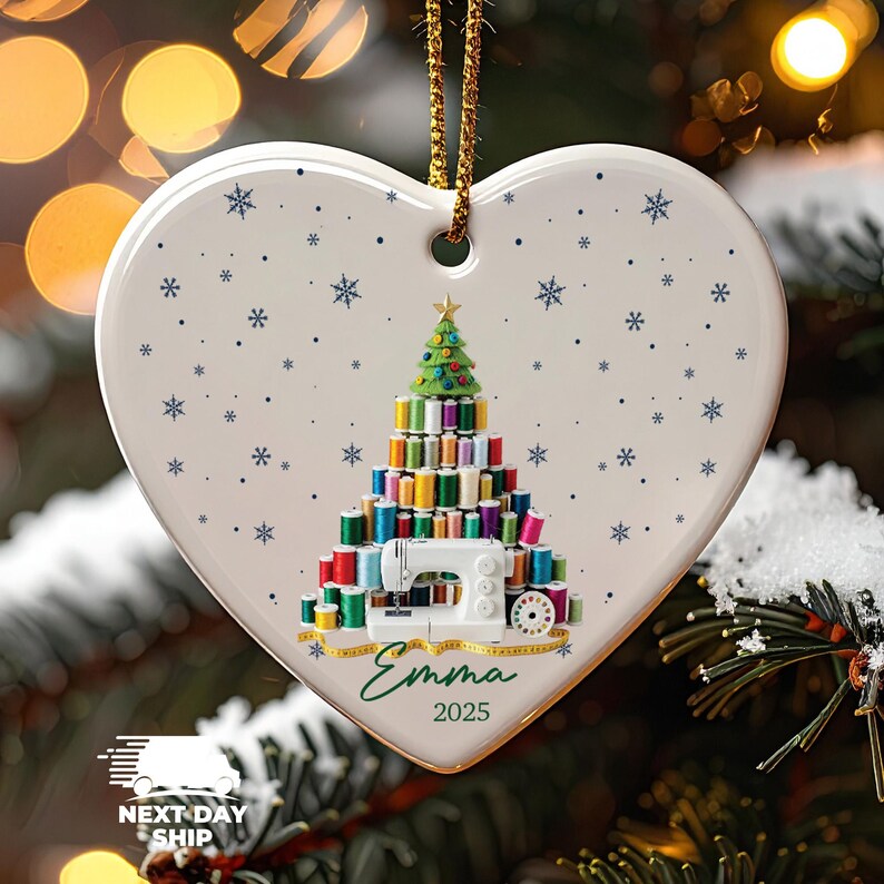 May include: A heart-shaped white ceramic ornament featuring a Christmas tree made of colorful sewing thread spools, a white sewing machine, and a yellow measuring tape. The design includes blue snowflakes, the name Emma, and the year 2025 in green text.