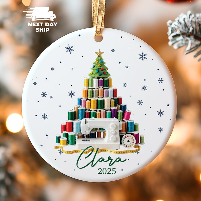 May include: A round white ceramic ornament featuring a Christmas tree constructed from colorful sewing thread spools, topped with a star. A white sewing machine sits at the base with a yellow measuring tape, the name Clara, and the year 2025 printed below.