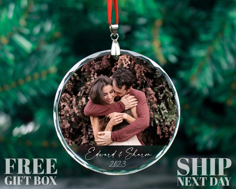 May include: A clear glass Christmas ornament with a photo of a couple hugging. The ornament has a silver trim and a red ribbon. The text "Edward & Sharon 2023" is printed on the ornament.
