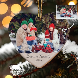 May include: A heart-shaped ceramic Christmas ornament featuring a printed photograph of a family of four in winter clothing holding gifts outdoors. The ornament includes a line-art Christmas tree graphic and the text Brown Family 2025.