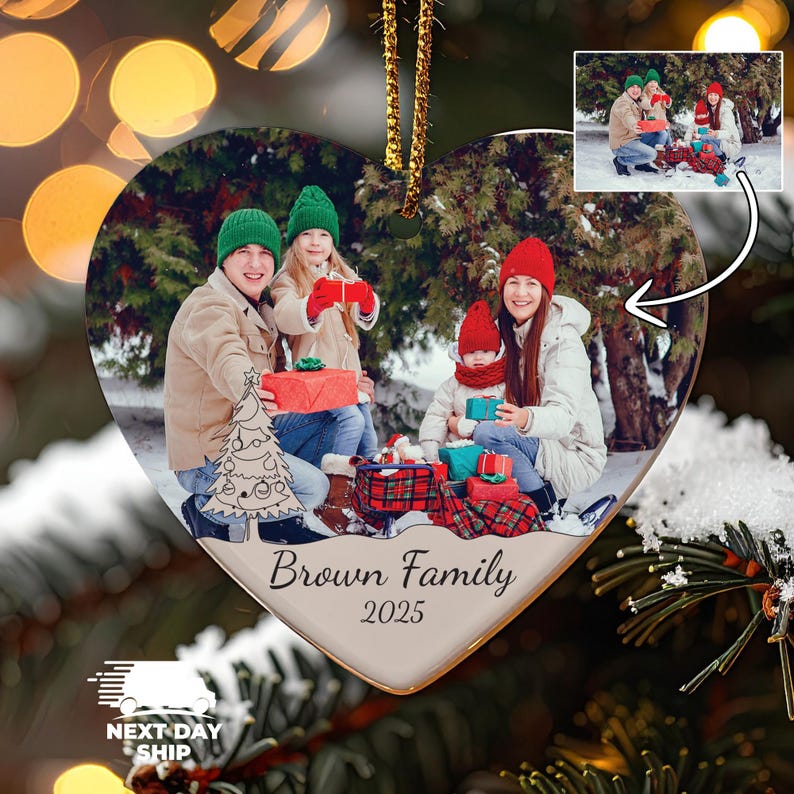 May include: A heart-shaped ceramic Christmas ornament featuring a printed photograph of a family of four in winter clothing holding gifts outdoors. The ornament includes a line-art Christmas tree graphic and the text Brown Family 2025.