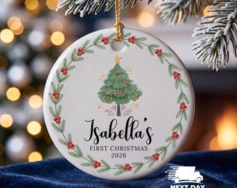 May include: A round white ceramic ornament featuring a green Christmas tree illustration with red ornaments and a gold star. A wreath of green leaves and red berries surrounds the tree. The text reads Isabella's FIRST CHRISTMAS 2026.