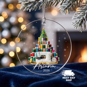 May include: A clear circular acrylic ornament featuring a printed design of a Christmas tree constructed from colorful sewing thread spools. A white sewing machine sits at the base with a yellow measuring tape. The text reads Ariana 2026.