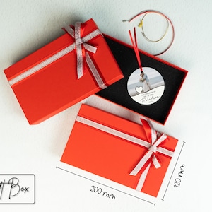May include: Red gift box with a silver ribbon and bow. The box is 8 inches by 12 inches. Inside the box is a round, white ornament with a photo of a couple and the text "First Christmas as Mr. & Mrs. Robertson 2023".