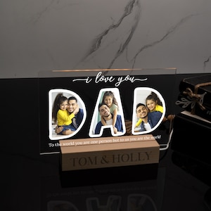 May include: A clear acrylic sign with the words "i love you DAD" in white lettering. The letters are filled with photos of a father and his children. The sign has a wooden base with the text "TOM & HOLLY" in white lettering. The sign reads "To the world you are one person but to us you are the world."