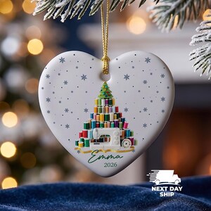 May include: A white heart-shaped ceramic ornament hanging from a gold string. The front features a graphic of a Christmas tree constructed from colorful sewing thread spools, a white sewing machine, the name Emma, and the year 2026 in blue text.