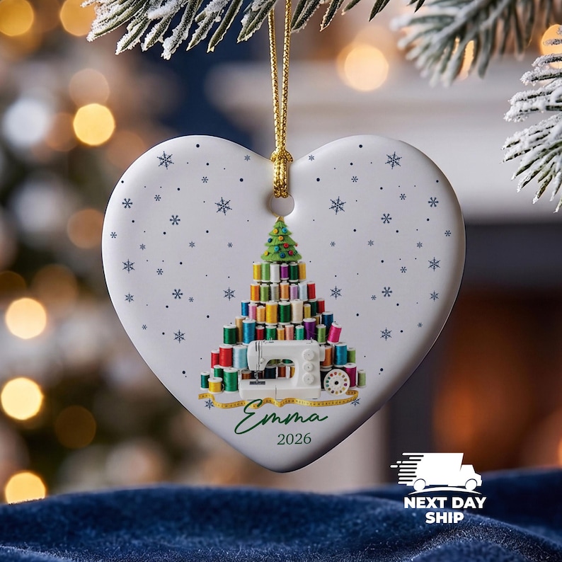 May include: A white heart-shaped ceramic ornament hanging from a gold string. The front features a graphic of a Christmas tree constructed from colorful sewing thread spools, a white sewing machine, the name Emma, and the year 2026 in blue text.