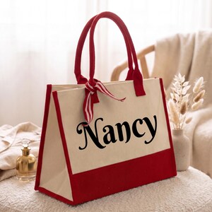 K&ouml;nnte beinhalten: Eine cremefarbene Tragetasche mit rotem Boden, roten Griffen und einem rot-wei&szlig; gestreiften Band, das an einem Griff befestigt ist. Der Name Nancy ist in gro&szlig;en, schwarzen Schreibschriftbuchstaben auf der Vorderseite der Tasche aufgedruckt.