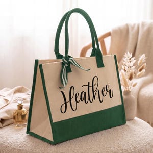 K&ouml;nnte beinhalten: Eine beigefarbene Tragetasche aus Segeltuch mit dunkelgr&uuml;nen R&auml;ndern und Griffen. Die Tasche ist auf der Vorderseite mit dem Namen Heather in schwarzer Schreibschrift versehen und hat ein gr&uuml;n-wei&szlig; gestreiftes Zierband an einem der Griffe.