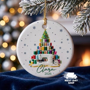 May include: A round white ceramic Christmas ornament featuring a graphic of a tree constructed from colorful sewing thread spools, topped with a green felt tree, and a white sewing machine at the base. The name Clara and the year 2026 are printed below.
