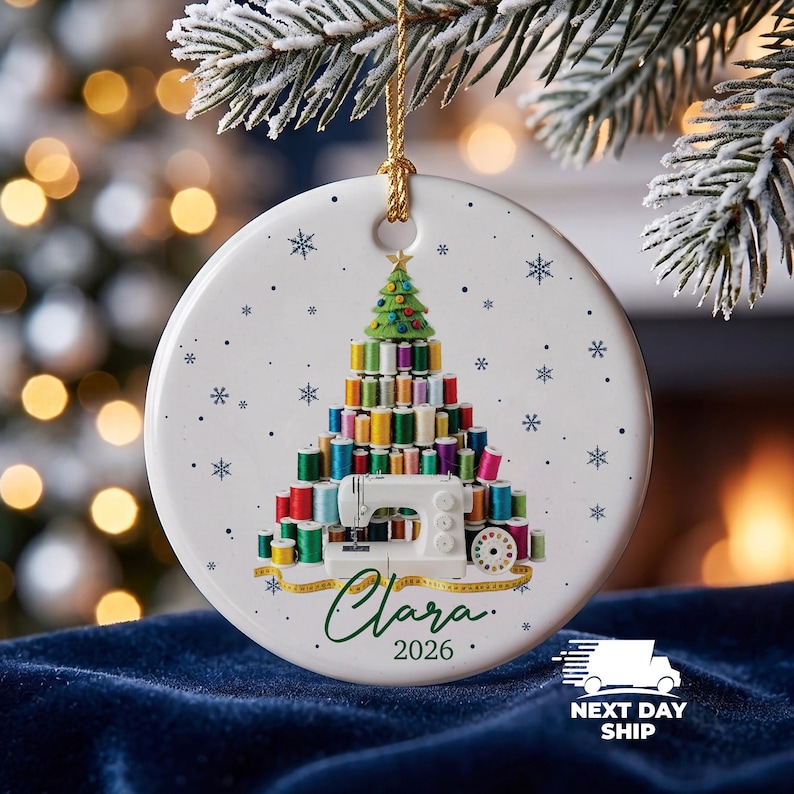 May include: A round white ceramic Christmas ornament featuring a graphic of a tree constructed from colorful sewing thread spools, topped with a green felt tree, and a white sewing machine at the base. The name Clara and the year 2026 are printed below.