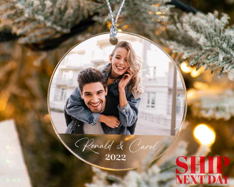 May include: A round, clear glass Christmas bauble featuring a photo of a couple. The woman is smiling and has her arm around the man's neck. The man is smiling and looking at the camera. The bauble has the text "Ronald & Carol 2022" printed on it.