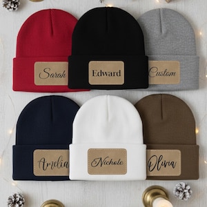 May include: A collection of six beanies in various colors: red, black, gray, navy blue, white, and brown. Each beanie has a rectangular patch with a name or the word "Custom" in a cursive font. The beanies are displayed on a light-colored surface.