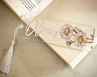 May include: A clear glass bookmark with a white tassel. The bookmark has the name "Olivia" written in white script and a floral design with white daisies and brown stems.