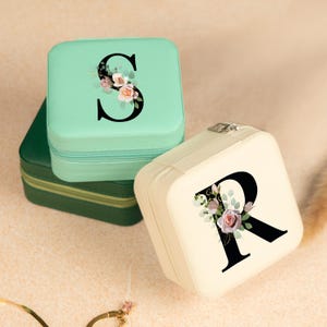 May include: Two small square jewelry boxes with floral designs and a single letter on each. The top box is light green with the letter "S" and the bottom box is cream with the letter "R".