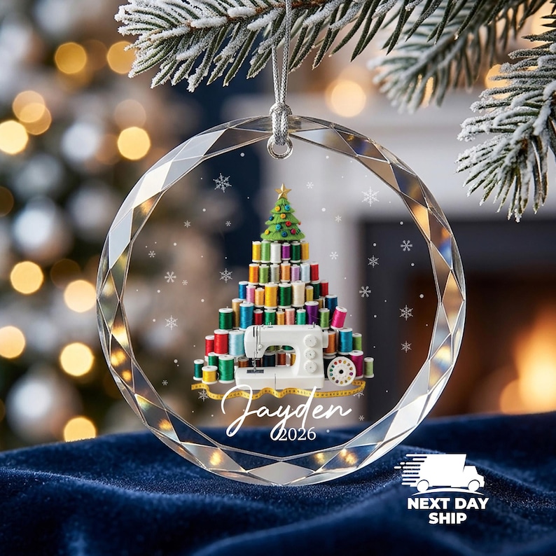 May include: A round, faceted clear glass ornament featuring a printed graphic of a Christmas tree constructed from colorful sewing thread spools, a white sewing machine, and a yellow measuring tape. The name Jayden and the year 2026 are printed below.