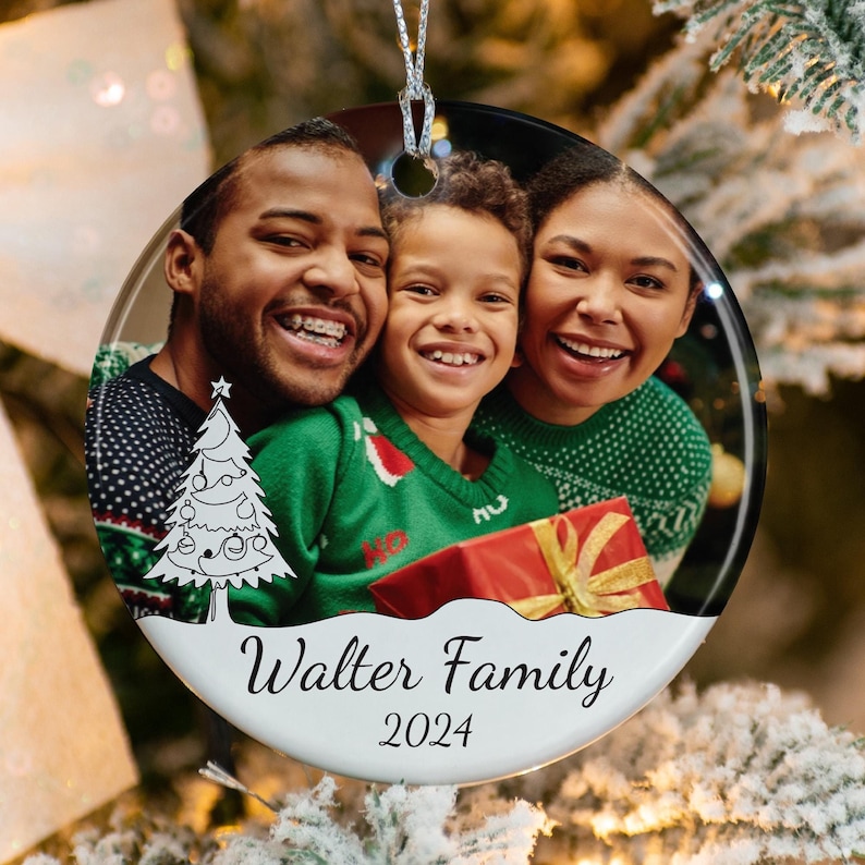 May include: A round ceramic Christmas ornament featuring a color photograph of a family of three smiling. The ornament includes a white base with a black line drawing of a Christmas tree, the text "Walter Family" and the year "2024" in black.