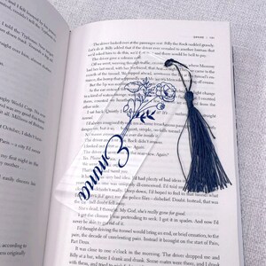 May include: A clear glass bookmark with a floral design and a blue tassel. The bookmark is resting on an open book with text about a tunnel.