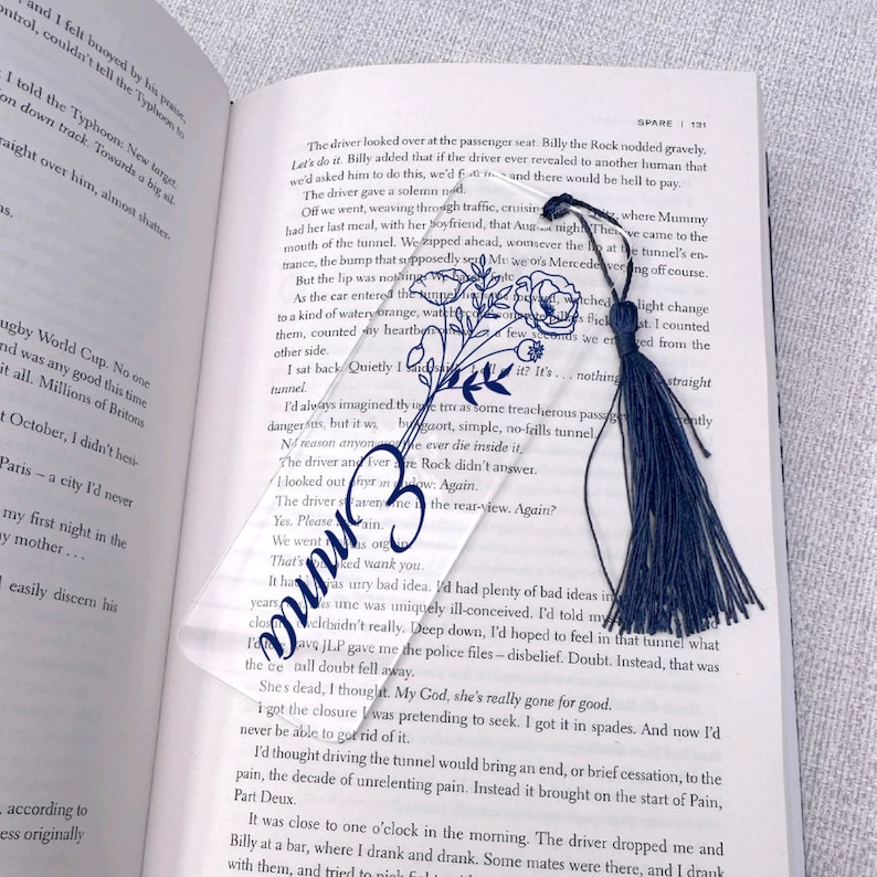 May include: A clear glass bookmark with a floral design and a blue tassel. The bookmark is resting on an open book with text about a tunnel.