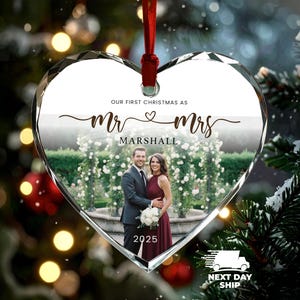 May include: Heart-shaped glass Christmas ornament with a photo of a couple. The ornament reads "OUR FIRST CHRISTMAS AS Mr & Mrs MARSHALL 2025". A red ribbon is attached for hanging. The background includes Christmas tree branches and lights.