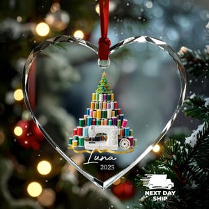 May include: A heart-shaped clear acrylic ornament featuring a printed graphic of a Christmas tree constructed from colorful sewing thread spools, topped with a star, and a white sewing machine at the base. The name Luna and year 2025 are printed below.