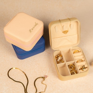 May include: A beige square jewelry box with a gold zipper and the name "Hannah" written on the top. The box is open and shows a compartment for rings and earrings. The box is sitting on a blue jewelry box.