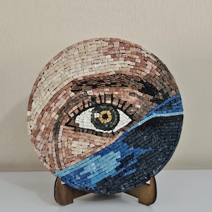 以下が含まれることがあります： 詳細な人間の目が描かれた円形のモザイク壁掛けアート。虹彩は緑と黄色で、白い強膜と暗い色のまつ毛に囲まれています。周囲の肌の色調はベージュとタンのタイルで構成されており、青と黒のマスクが施されています。