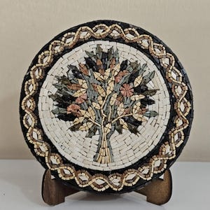 May include: A circular stone mosaic plaque featuring a central tree design with green, tan, and pink foliage. The border consists of a black and tan interwoven knot pattern. The mosaic is displayed on a small, brown, two-piece wooden stand.