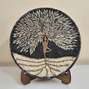 Könnte beinhalten: Ein rundes Mosaik-Wandbild mit einer stilisierten menschlichen Figur in Yoga-Pose, die in einen Baumstamm integriert ist. Der Baum hat hellgrüne Steinblätter, einen braunen Stamm und weiße Kieselwurzeln vor einem schwarzen Hintergrund.