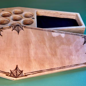 May include: A wooden box with a coffin shape and a black felt interior. The lid is decorated with a spiderweb design. The box has a compartment with six round holes.
