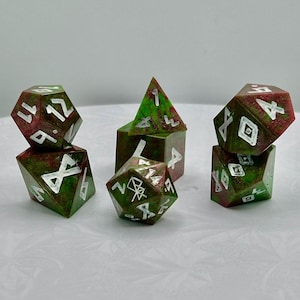 May include: A set of polyhedral dice in various shapes, including a pyramid, a cube, and other multi-sided dice. The dice are a mix of green and maroon with white numbering and symbols. The dice are designed for tabletop gaming.
