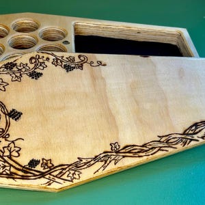 May include: A wooden box with a hinged lid. The lid is decorated with a wood-burned design of vines and leaves. The box has a compartment with multiple circular cutouts.