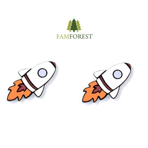 May include: Two white rocket ship shaped wooden ornaments with orange flames. The rockets have a red stripe and a white circle in the middle.