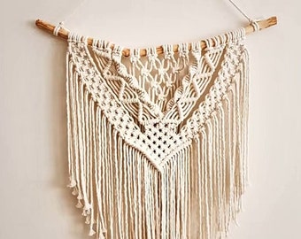 May include: A white macrame wall hanging with a geometric pattern and fringe. The hanging is attached to a natural wood branch.
