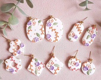 May include: A collection of white floral earrings with gold-colored posts. The earrings feature various shapes, including ovals, teardrops, and circles, adorned with purple, pink, and green floral designs. The earrings are set against a soft pink background.
