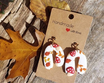 May include: A pair of white U-shaped dangle earrings featuring a pattern of red mushrooms with white spots and orange autumn leaves. The earrings are attached to a rectangular brown cardboard display card with the text Handmade with love.