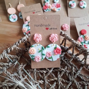 May include: A pair of round white earrings featuring raised pink and peach floral designs with green leaves and small yellow dots. The earrings are attached to a brown cardboard display card with the text Handmade with love printed on it.